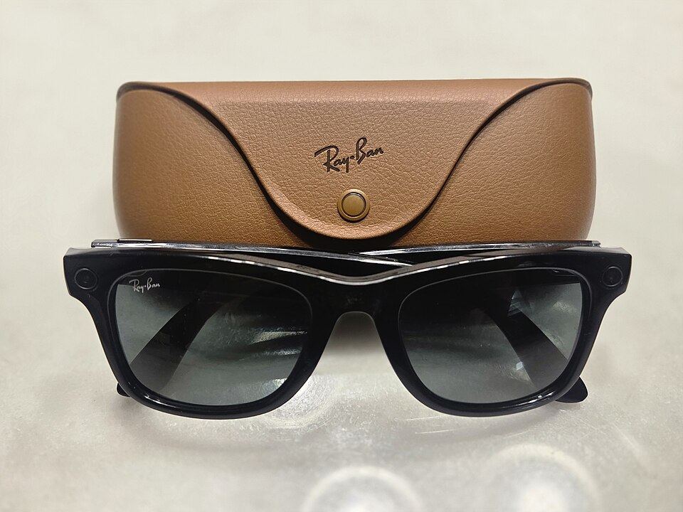 A pair of shiny black Ray-Ban Meta Gen 1 smart glasses positioned in front of their brown leather charging case. (Image Source: Wikimedia Commons | CCadio)