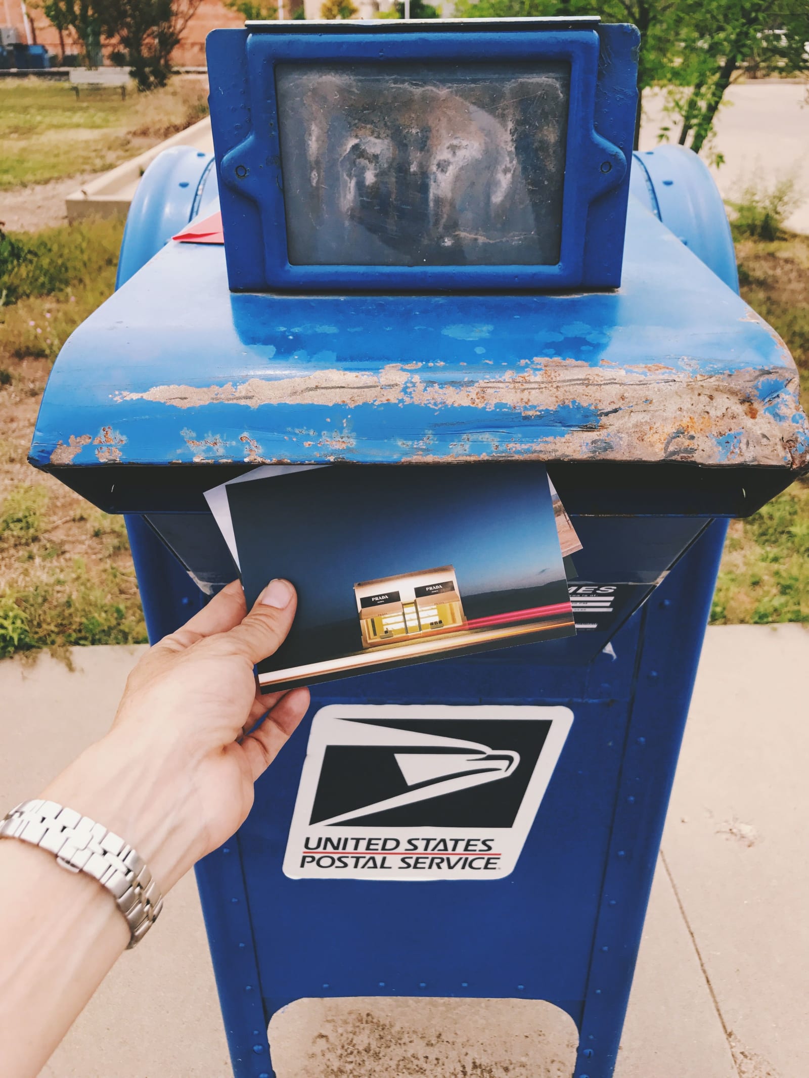 Close-up of a hand posting a postcard into a blue USPS mailbox.