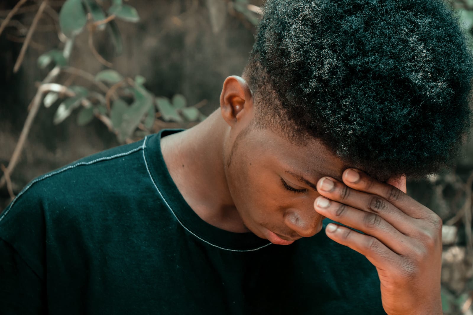 A young man with closed eyes and hand on forehead stands thoughtfully amidst greenery. (Representative Cover Image Source: Pexels | Photo by Derick Makwasi)