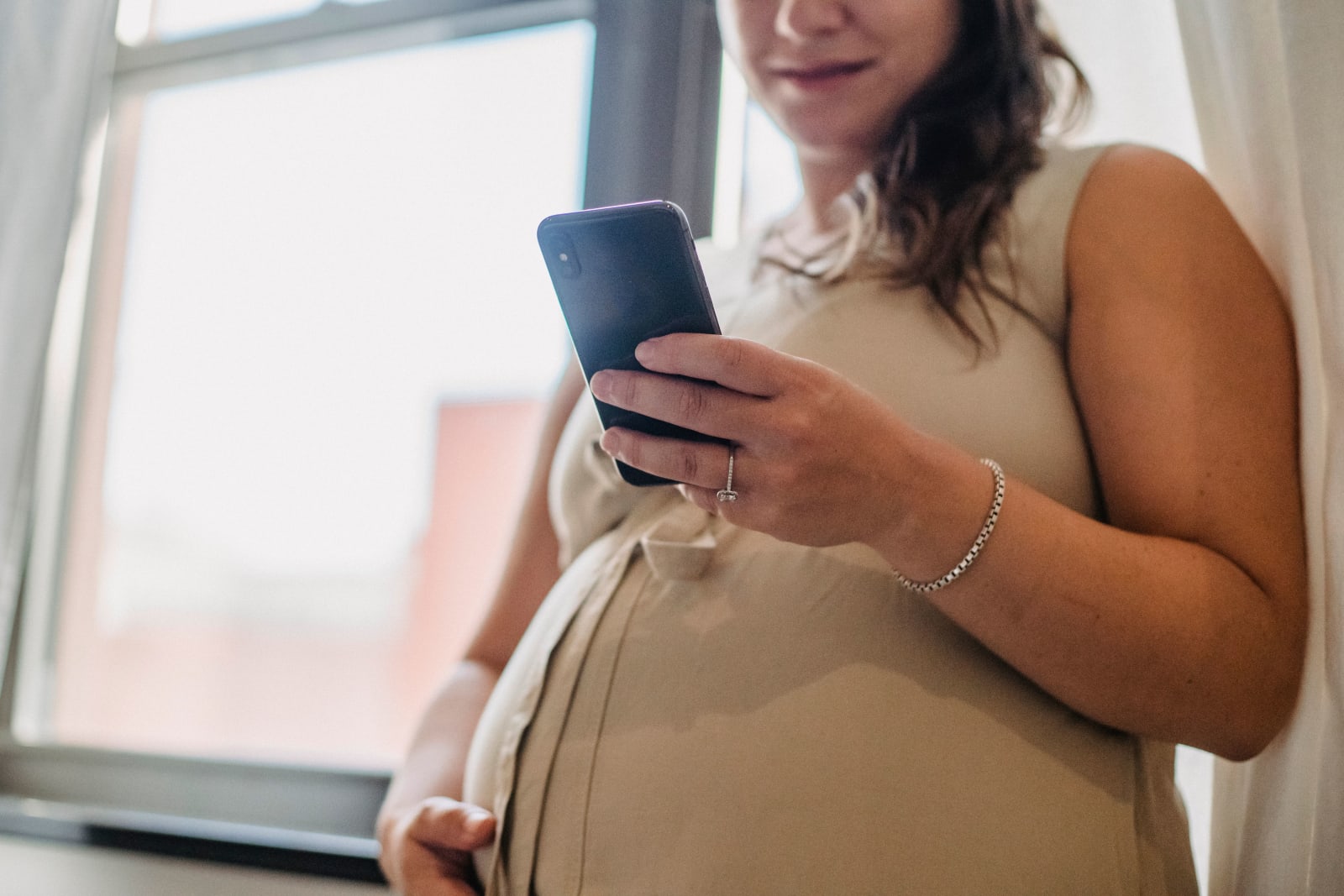 A pregnant woman stands by a window using her smartphone, enjoying a quiet moment indoors. (Representative Image Source: Pexels | Photo by Amina Filkins)