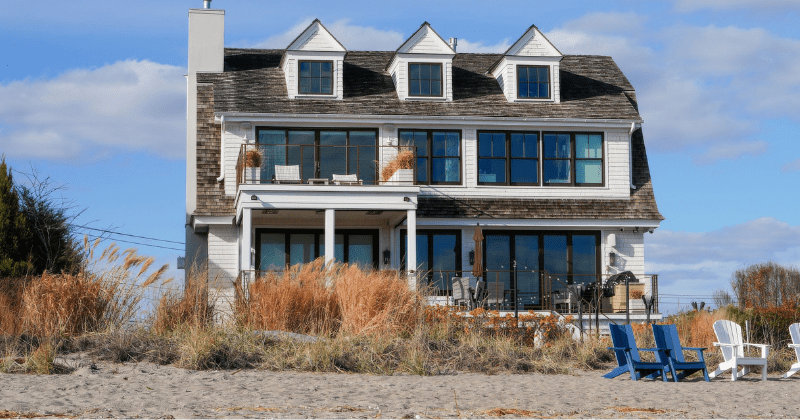 Cozy beachfront house with patio chairs surrounded by sand and grasses under blue sky (Representative Image Source: Pexels | Photo by Cara Denison)