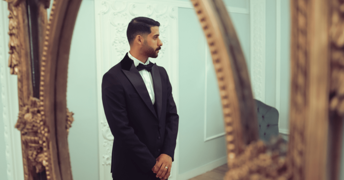 A sophisticated man in a tuxedo stands by an ornate mirror in an elegant room