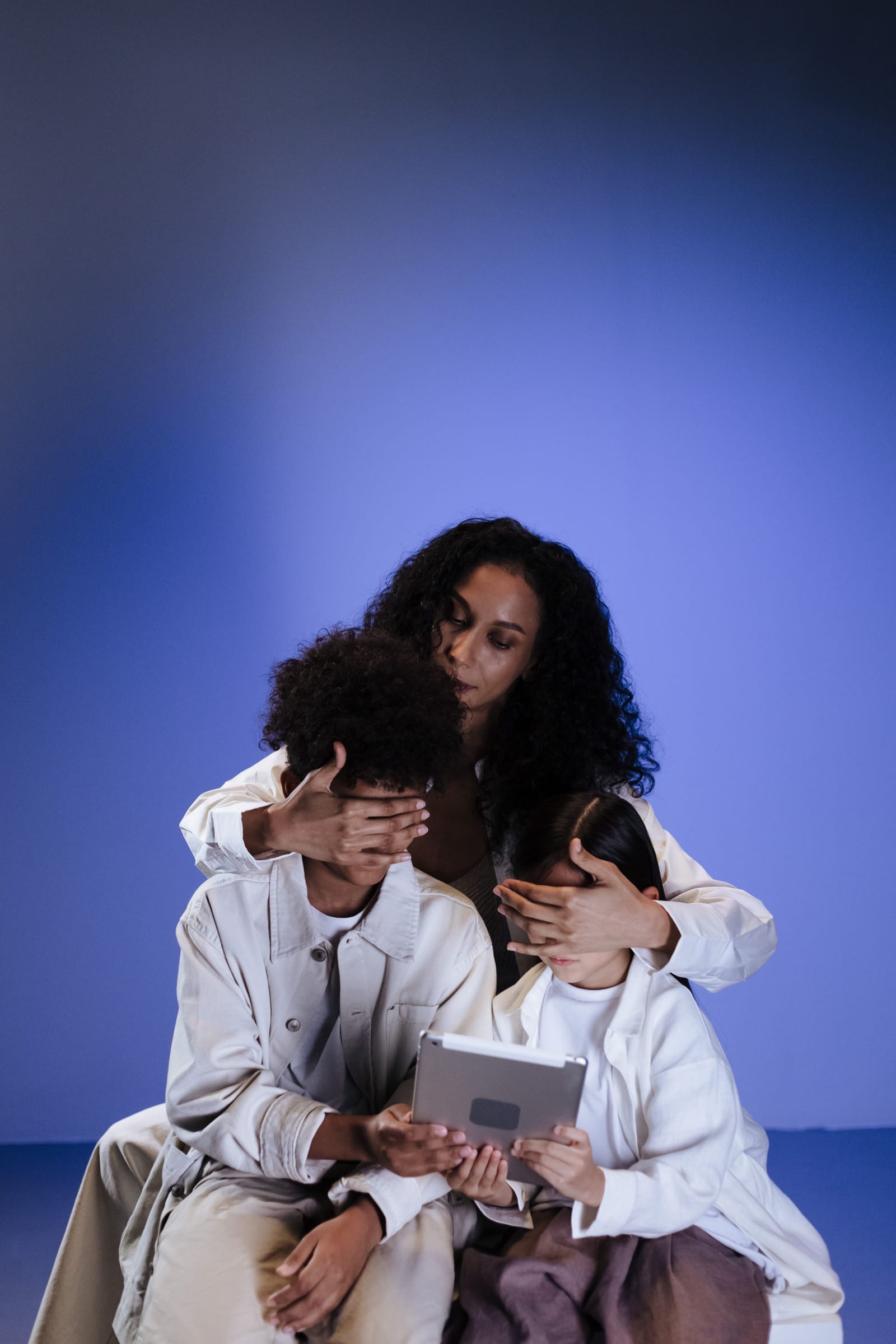 A mother uses her hands to cover her children's eyes while they sit with a tablet, highlighting safety concerns.
